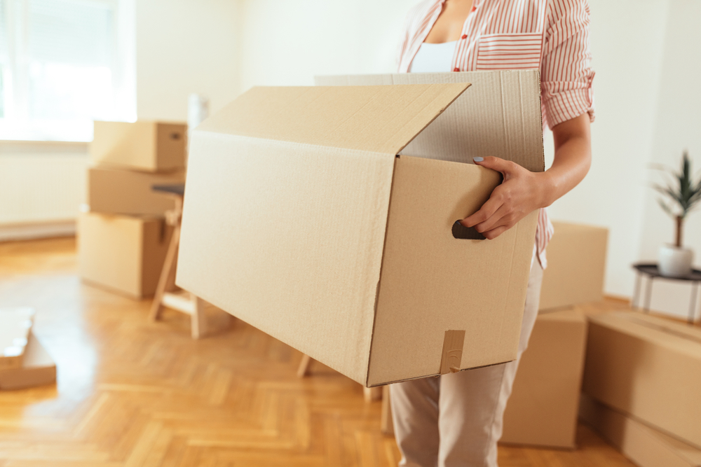 A women holding the handles of a moving box in a room with other moving boxes. 