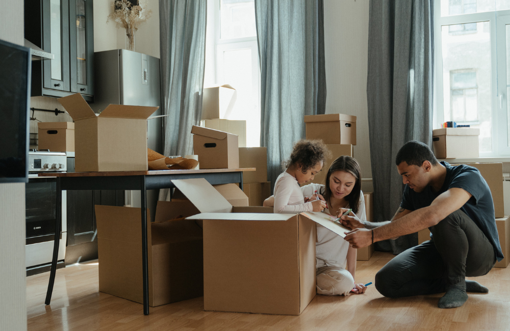 A mother, father and daughter in a kitchen filled with moving boxes. Their daughter is in an empty box and the father is drawing on the box.