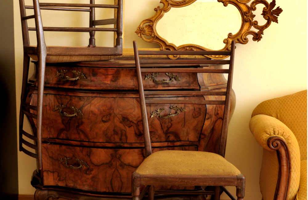 An antique set of drawers with an antique mirror on top and antique chairs stacked around it.