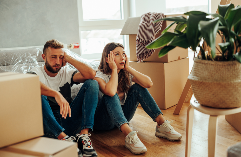 A couple sitting with moving boxes around them looking exasperated. 