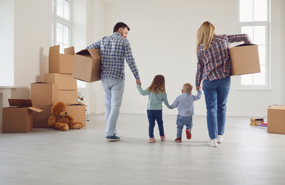 A mother and father walking in a mostly empty room with a amoving box under one arm and holding the hand of their son and daughter between them. There are a few moving boxes stacked in the room. 