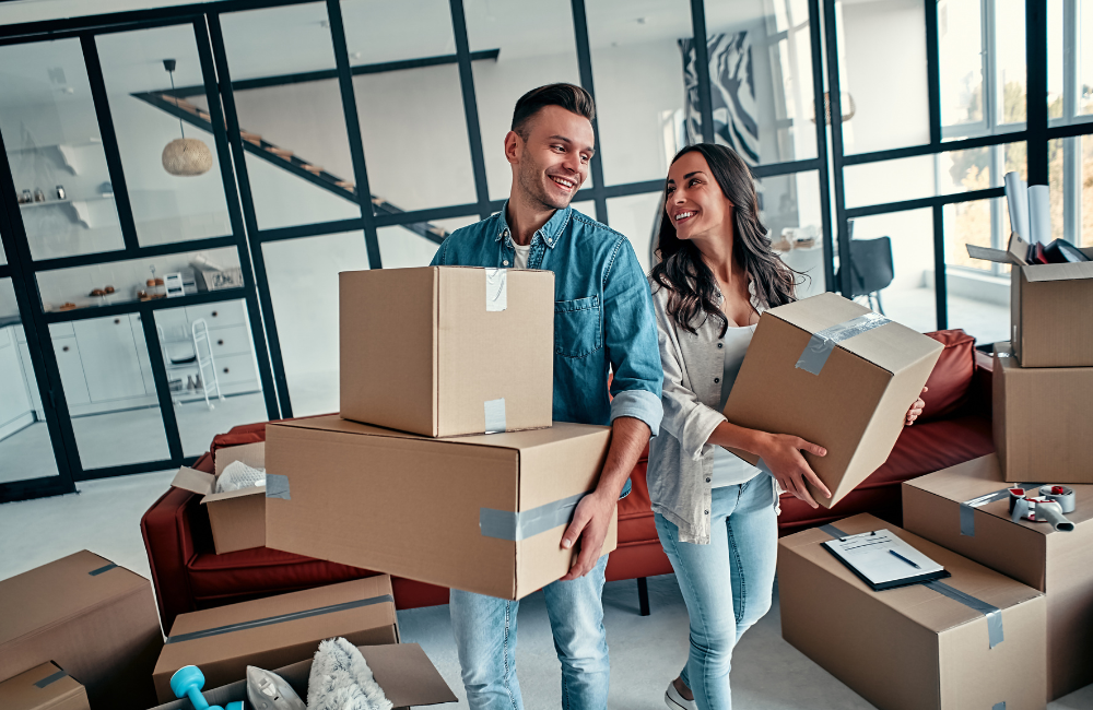 A couple ion a lounge room filled with moving boxes. The couple are smiling at each other. 