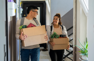A male and female smiling at each other both holding open moving boxes containing various items and plants.