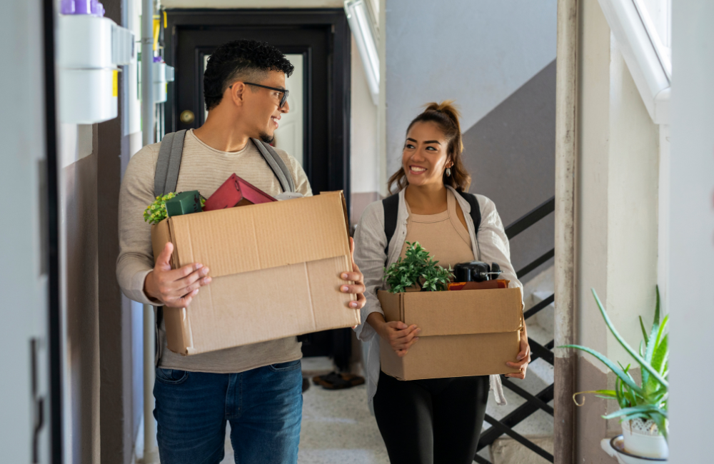 A male and female smiling at each other both holding open moving boxes containing various items and plants. 