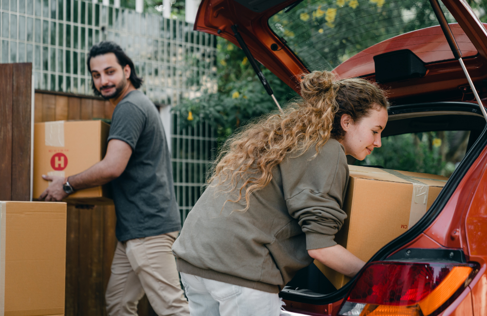 A woman putting a box into the boot of a car while a man watches also holding a moving box.