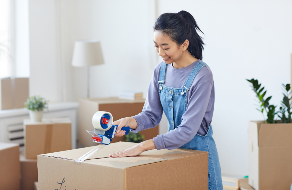 A girl in a lounge room filled with moving boxes. She is taping one of the boxes closed.