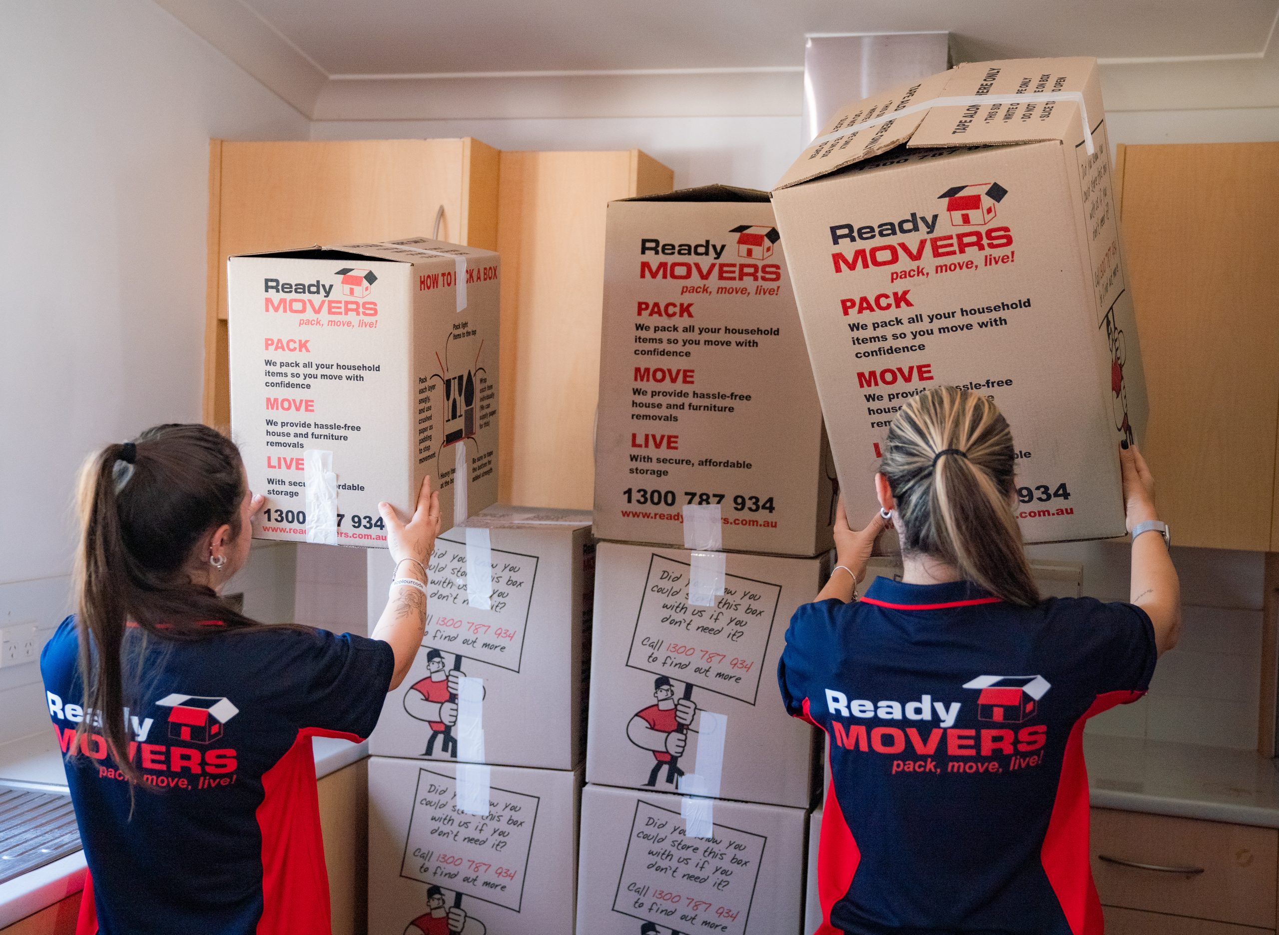 Two women wearing the Ready Movers' uniform stacking boxes against a wall. 