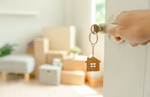 A door opening to a room filled with moving boxes. There is a key in the lock of the door with a mini wooden house as a keyring.