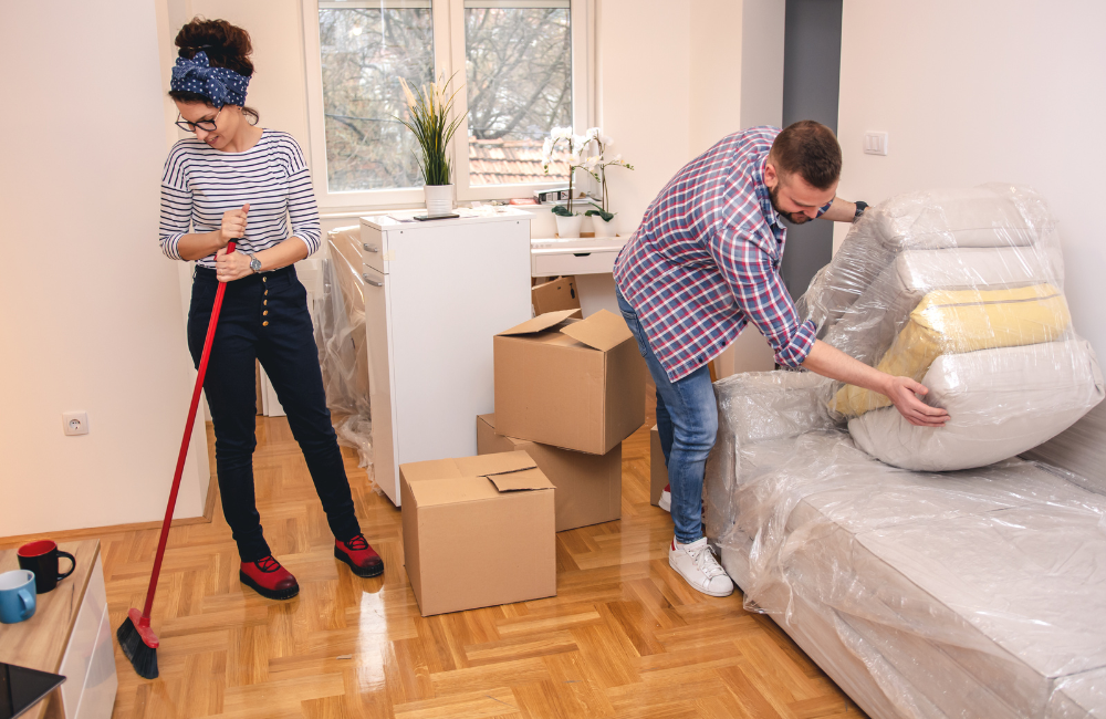 A couple in a house with all their belonging packed up ready to move. The woman is holding a broom while the man is stacking couch cushions wrapped in plastic. 