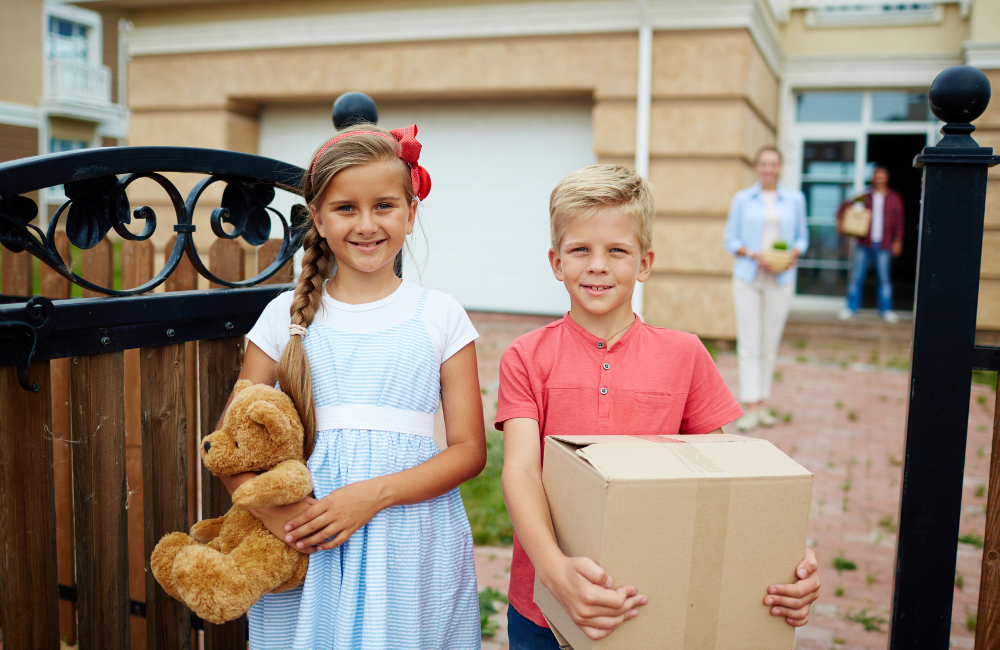 A boy and a girl standing at the front gate. The girl is holding a teddy bear and the boy is holding a small moving box. Their parents are behind them with moving boxes.