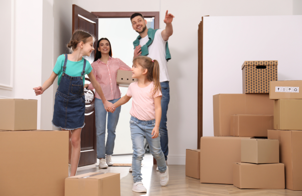 Parents and their two daughter walking into their new home carrying moving boxes. They all look happy and the father is pointing to something off camera.