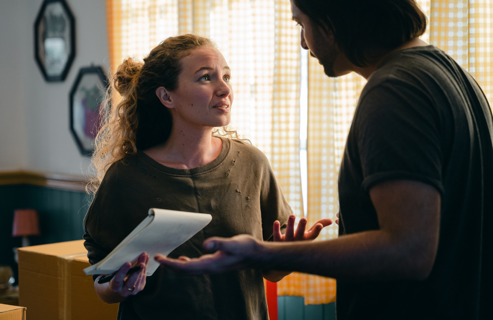 A couple having a disagreement with moving boxes around them. The woman is holding a notepad.