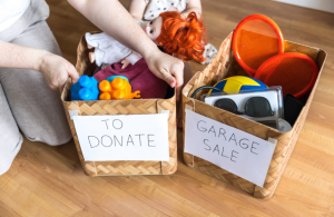 A person kneeling next to two baskets. One basket is labelled "To Donate" and the other reads "Garage Sale." The person is holding onto the side of the one labelled "To Donate."