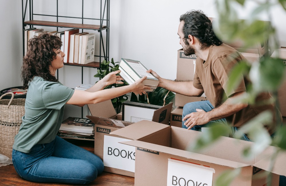 A couple surrounded by boxes. The woman is passing the man a pile of books.