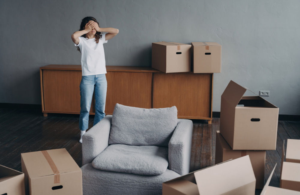 A woman standing in her lounge room with various moving boxes and furniture around her. Her hands are covering her face expressing stress. 