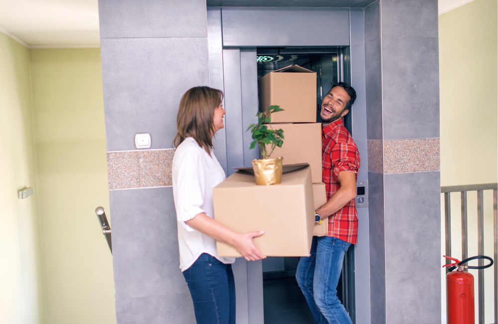 A woman and a man holding boxes in front of an elevator. The man is attempting to squeeze through the elevator doors. The people are laughing.