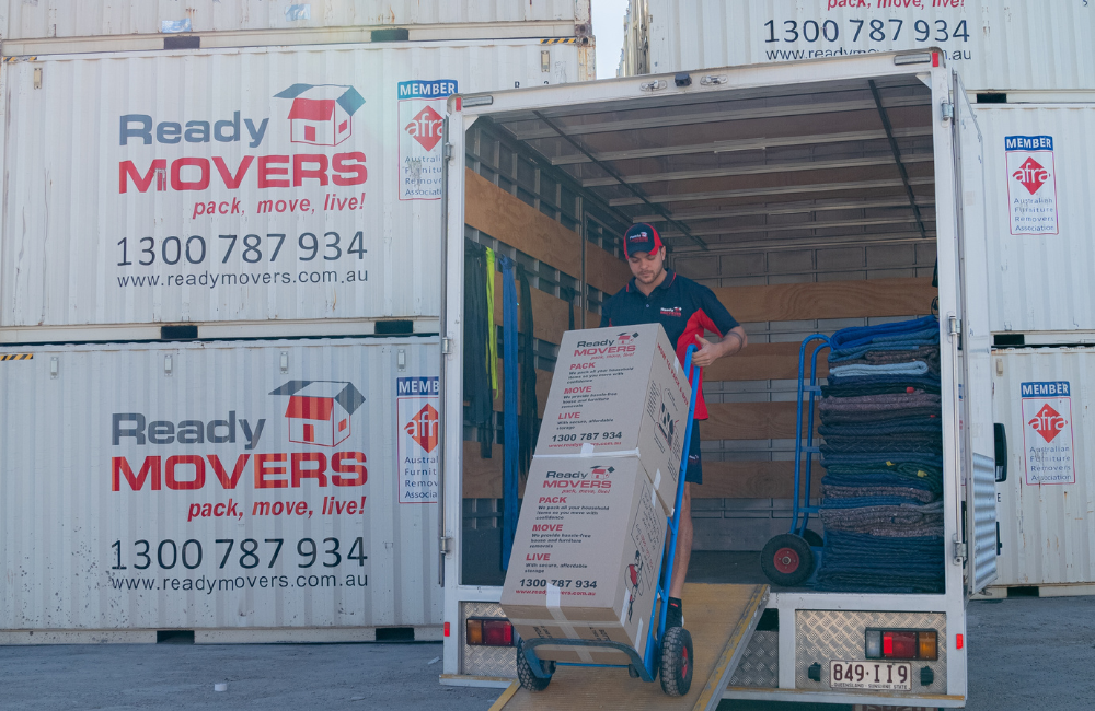 A Ready Movers removalist pulling a moving dolly with two boxes on it into a truck. There are shipping containers saying 'Ready Movers' in the background. 