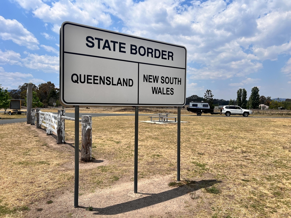 A state border signs indicating the line between Queensland and NSW. There is a grass surrounding it and a 4WD towing a caravan in the background.