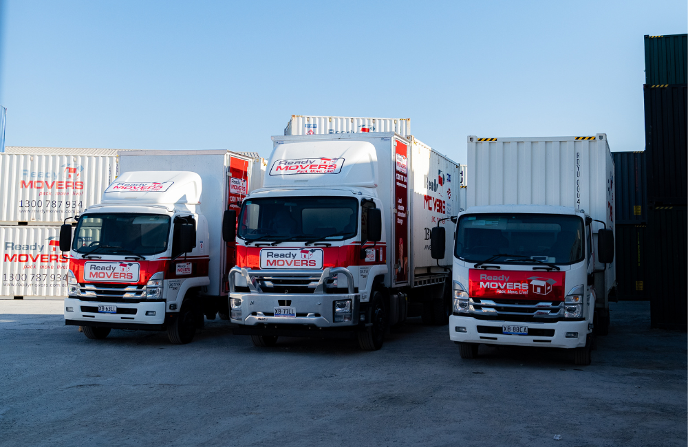 Three different Ready Movers removalist trucks lined up at a shipping yard with shipping containers in the background.