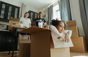 A family surrounded by boxes in their kitchen. Their daughter is sitting in one of the boxes while the mother looks on smiling.